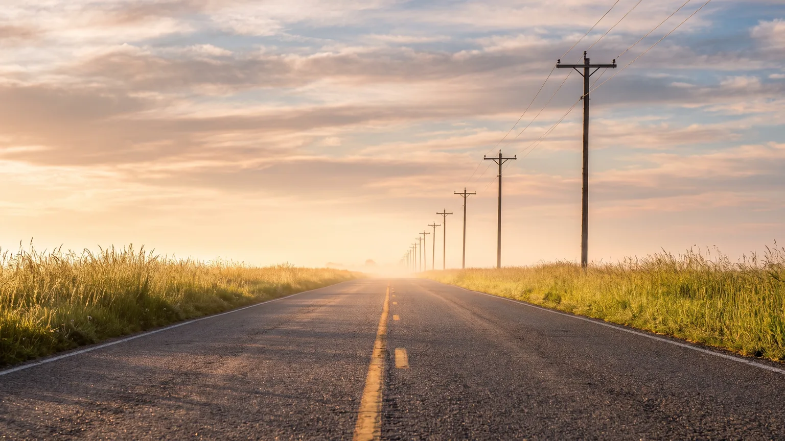 An empty two-lane highway running straight toward the horizon at sunrise, flanked by open grassland.