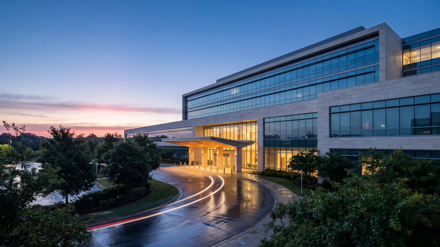 A modern medical centre at dawn, its lobby glowing warm against a deep blue pre-sunrise sky.